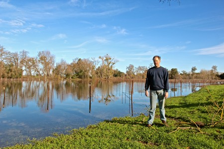 Man standing by a vineyard pond in Lodi, California, with clear blue sky and trees in the background.