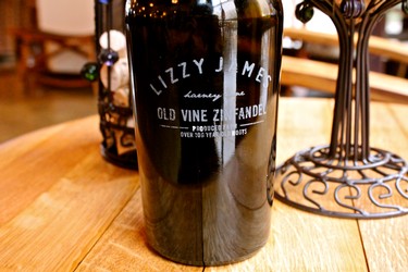 Bottle of Lizzy Meyer Old Vine Zinfandel wine on a wooden table, with decorative wine accessories in the background.