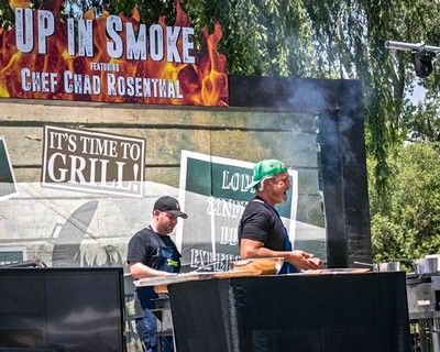 Two men grilling at a Lodi Winegrape Commission event with banners and trees in the background.