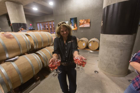 Woman holding glasses of rosé wine in a wine cellar with barrels in the background.