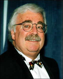 Close-up of a man with white hair, glasses, and a bow tie, smiling at the camera.