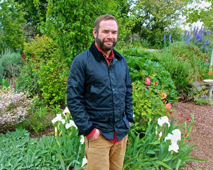 Man smiling in a garden with colorful flowers and lush green plants, wearing a navy jacket and khaki pants.