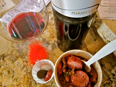 Glass of red wine, bottle, and bowl of sausage stew on a kitchen counter.