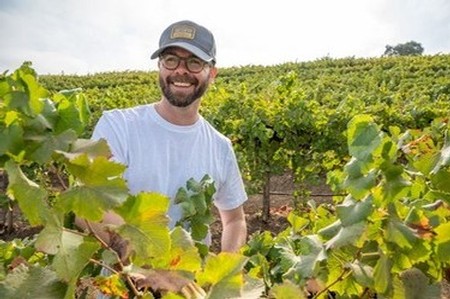 Happy vineyard worker in Lodi winegrape vineyard with lush green vines and rolling hills in the background.