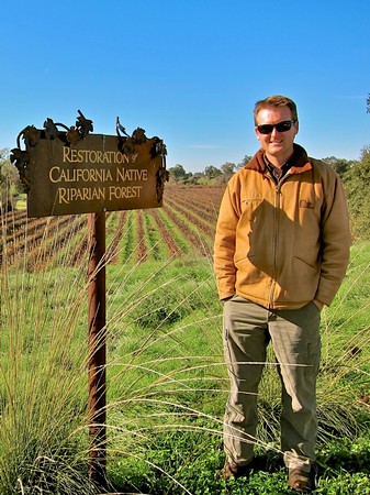 Man standing in a vineyard near a sign for California Native Riparian Forest restoration project.