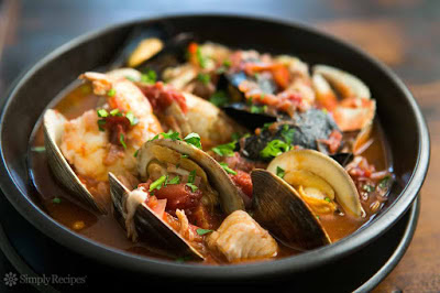 A bowl of seafood stew with clams, fish, and vegetables in a savory broth, served in a black bowl.