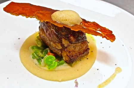 Elegant gourmet beef dish with crispy bacon, green vegetables, and creamy sauce on a white plate.