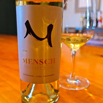Close-up of a 2016 Mensch wine bottle from Lodi, California, with a glass of white wine in the background.