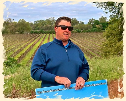 Man in sunglasses holding a sign in a vineyard with rows of grapevines behind him.