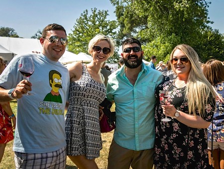 Group of people enjoying a sunny outdoor event with wine, smiling and holding glasses, surrounded by trees and tents.