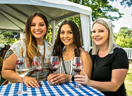 Three women smiling and holding wine glasses at an outdoor wine tasting event.