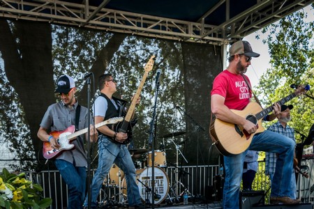 Band performing live on stage at Lodi Wine event with guitars and drums under trees.