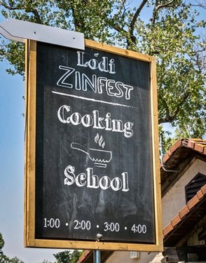 Lodi Zinfest cooking school sign with schedule, outdoor setting, trees, and building in background.