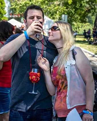 Two people enjoying wine at an outdoor event, with trees and other attendees in the background.