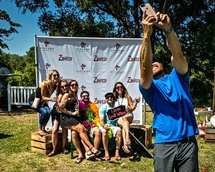 Group of people taking a selfie at Zinfest event with a backdrop and trees in the background.