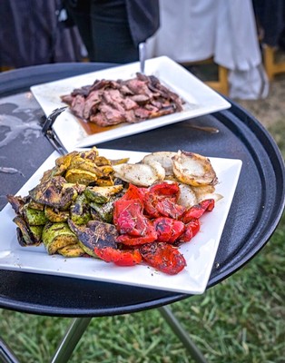 Variety of grilled vegetables and sliced meat served on white plates at an outdoor event.
