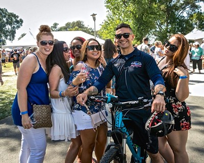 Group of smiling young adults enjoying outdoor wine event in Lodi, California, with a man on a bicycle and others holding up.