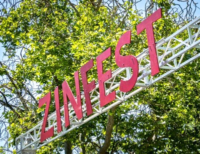Large pink ZINFEST sign on a white metal structure among green trees.