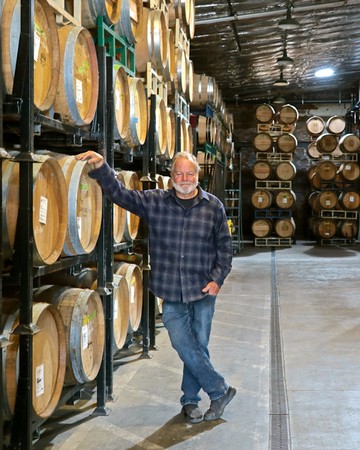 Man in winery cellar with wine barrels, smiling and pointing at barrels.