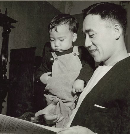Black and white vintage photo of a father and child reading a book together at home.