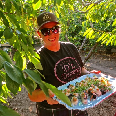 Woman holding a tray of sushi outdoors among green leaves, smiling at the camera.