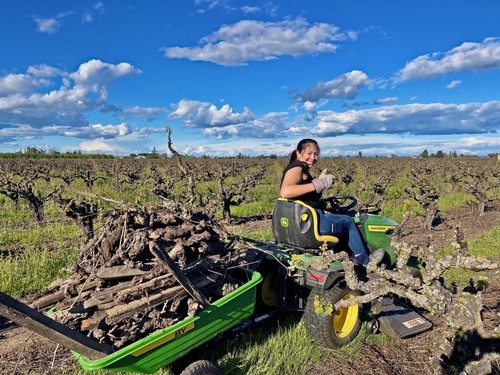 Woman operating a tractor in a lush vineyard during daytime with blue sky and clouds.