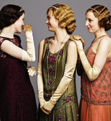 Three women in vintage dresses and gloves smiling and talking, styled in 1920s fashion.