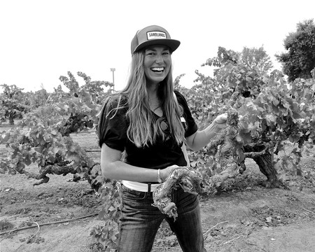 Happy woman in vineyard with grapevines, smiling, wearing a cap and casual clothes, during grape harvest.