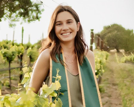 Smiling woman in vineyard wearing a teal sash, celebrating Lodi winegrape harvest.