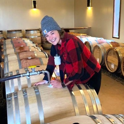 Young woman in a beanie and red plaid shirt smiling at the camera in a wine cellar with barrels.