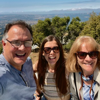 Group of three smiling people outdoors with vineyards and mountains in the background.