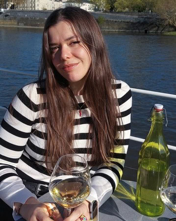Woman with long brown hair in a striped shirt holding a glass of white wine by the river during daytime.
