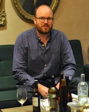 Man with glasses and beard at wine tasting event, surrounded by wine bottles and glasses.