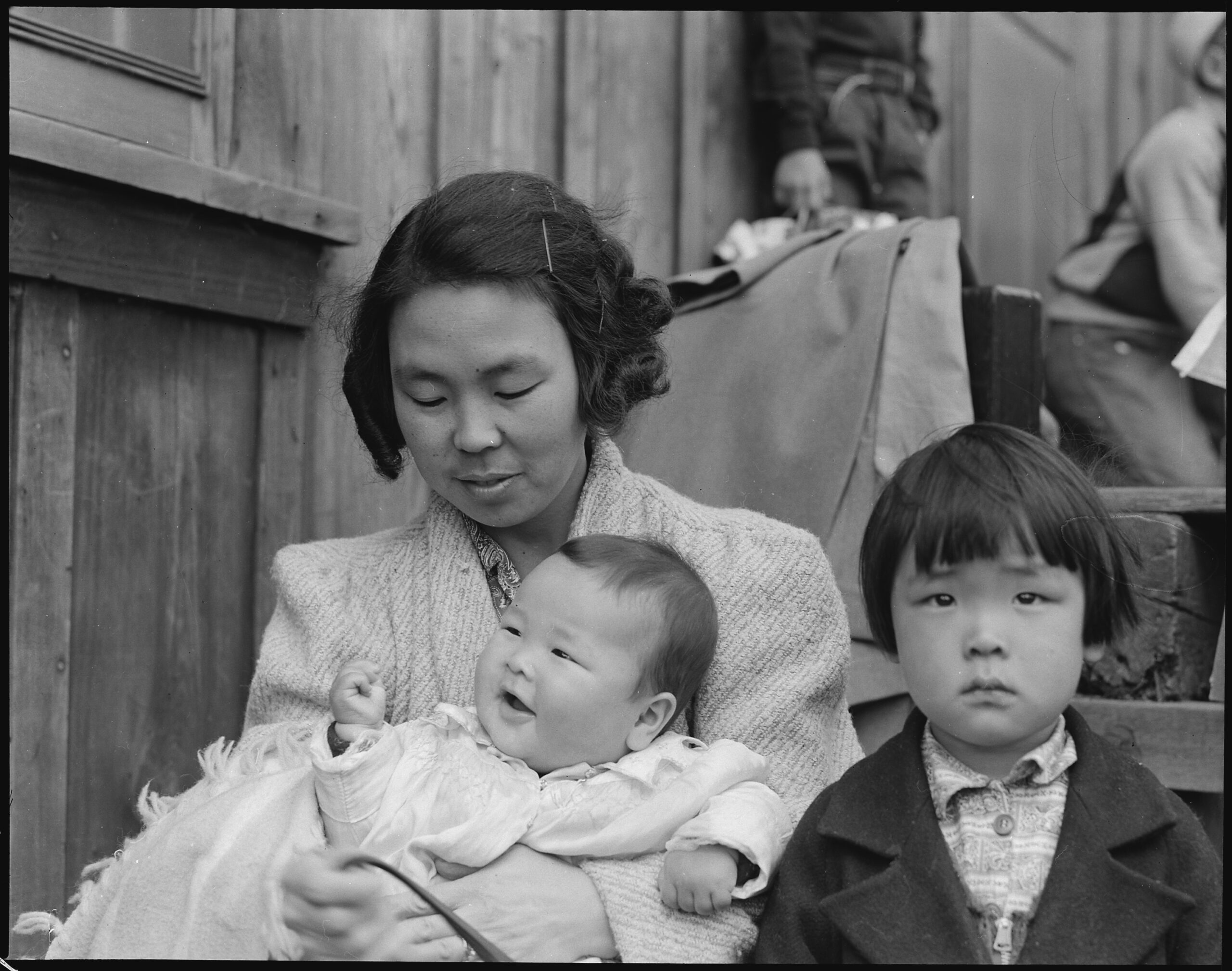 Historical black and white photo of a woman with two children, one infant and one young boy, in a rustic outdoor setting.