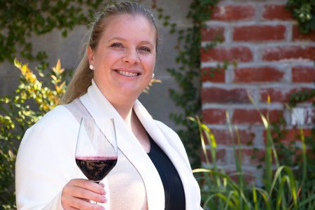 Woman holding a glass of red wine outdoors, smiling, with a garden background.
