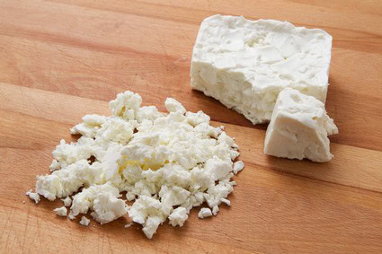 Crumbled white cheese with a block on a wooden cutting board, close-up view.
