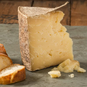 Close-up of a wedge of aged cheese with a rustic rind and crumbly texture, with bread slices nearby.