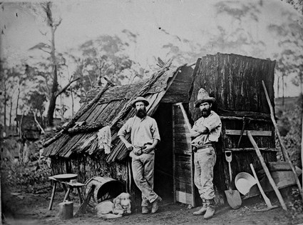 Two men standing outside a rustic, weathered wine cellar in Lodi, California, with vintage tools and a dog nearby.