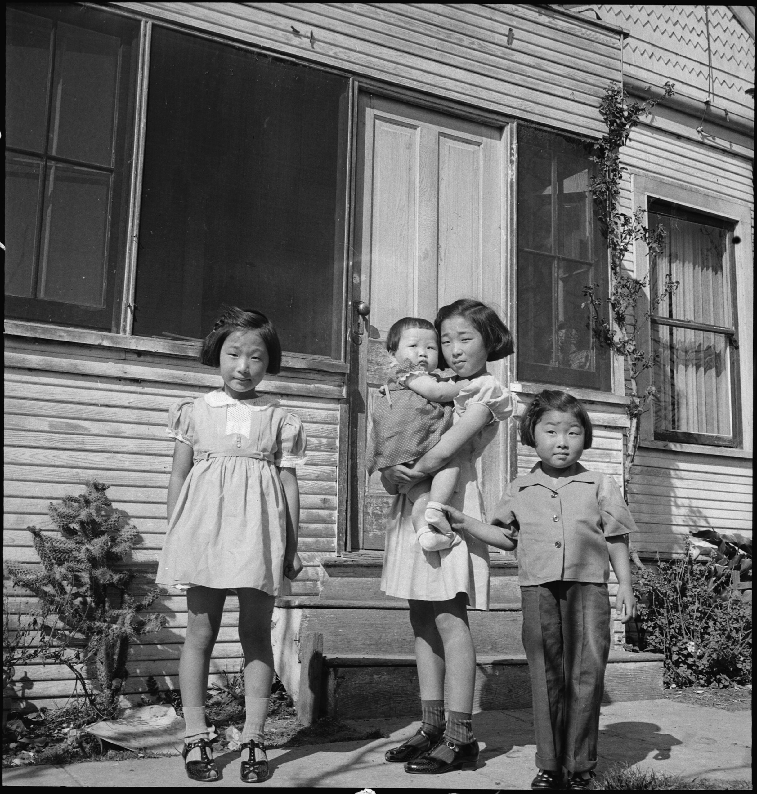 Historical black and white photo of four children standing outside a wooden house.
