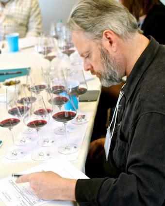 Man evaluating wine samples at a tasting event with multiple glasses of red wine.