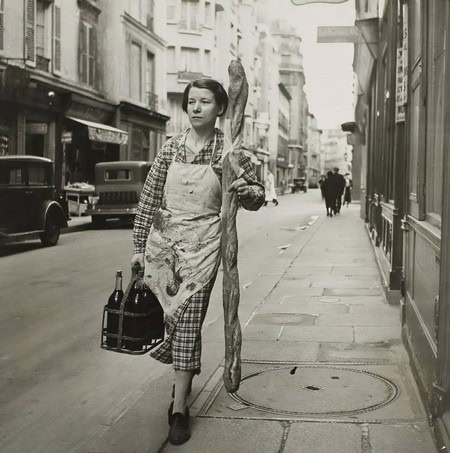 Black and white photo of a woman walking on a city street carrying a baguette and a basket, vintage urban scene.
