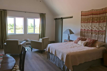 Bright bedroom with large window, comfortable chairs, and a bed with decorative pillows and a tapestry.
