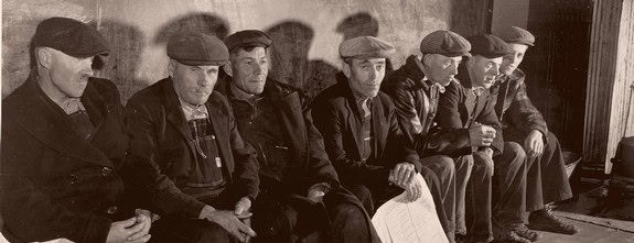 Black and white photo of men sitting in a row, wearing caps and jackets, in a vintage setting.