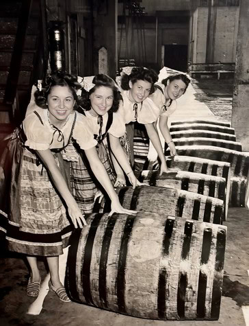 Four women in vintage dresses working with wine barrels in a winery setting.