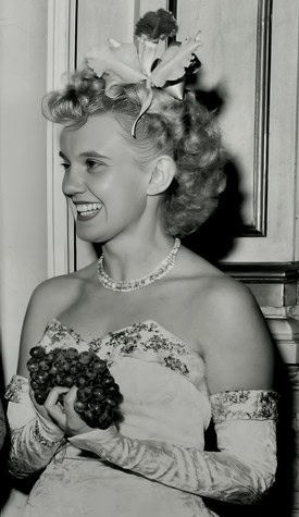 Black and white vintage photo of a woman holding grapes, wearing a floral dress and a pearl necklace.
