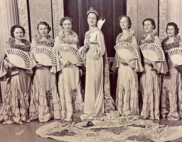 Group of women in traditional dresses with fans, standing with a crowned woman in the center, vintage photo.