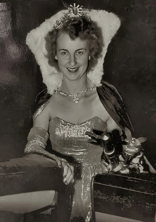 Black and white photo of a woman in a fancy dress with a tiara and jewelry, smiling at the camera.
