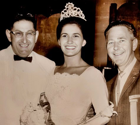 Young woman wearing a tiara at a celebration with two men, all smiling, in a vintage-style photo.