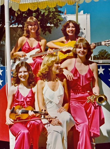 Group of five women in pink and white dresses playing guitars and saxophone on stage at an outdoor event.