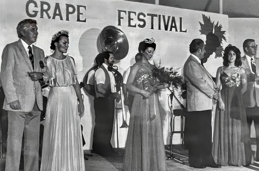Historical black and white photo of people at a grape festival, with women in elegant dresses and men in suits, on stage.